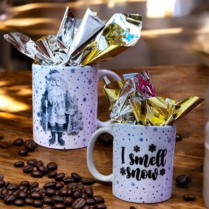 Peut inclure: Deux tasses en céramique blanche avec des motifs de flocons de neige bleus. Une tasse présente une illustration de Père Noël et l'autre tasse dit "Je sens la neige".