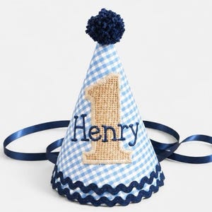 May include: A conical party hat featuring a light blue and white gingham pattern. The hat is topped with a dark blue pom-pom and trimmed with dark blue rickrack. A burlap number one is appliqued on the front with the name Henry embroidered in dark blue thread. Dark blue satin ribbons are attached to the sides.