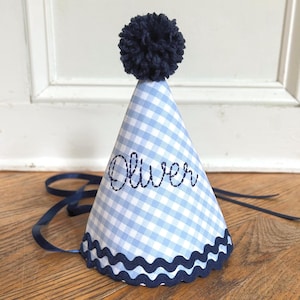 May include: A conical party hat featuring a light blue and white gingham pattern. The hat is topped with a dark navy blue yarn pom-pom, trimmed with a dark navy blue wavy rickrack border, and personalized with the name Oliver in dark blue script.