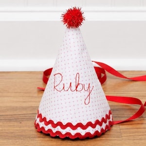 May include: A white cone-shaped party hat featuring a pattern of small red dots, the name Ruby embroidered in red script, a red pom-pom at the top, a red wavy trim along the bottom edge, and long red satin ribbons for securing the hat in place.