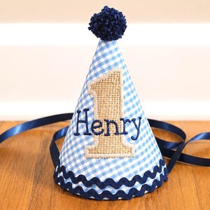 May include: A conical party hat featuring a light blue and white gingham pattern, topped with a dark blue pom-pom. The front displays a hessian number one with the name Henry embroidered in dark blue thread, finished with a dark blue wavy trim.