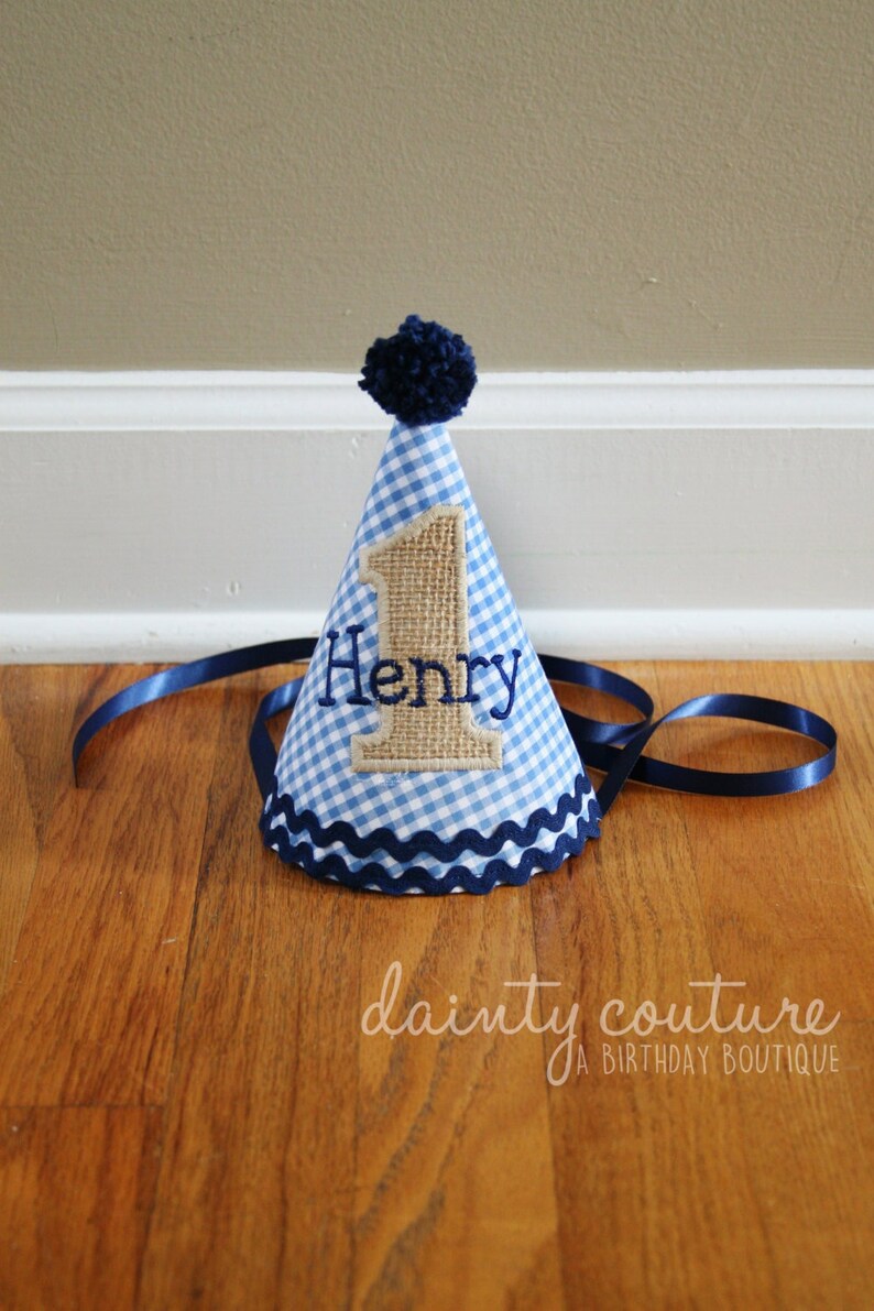 May include: A cone-shaped party hat featuring a light blue and white gingham pattern, topped with a dark blue pom-pom. The hat displays a burlap-textured number one with the name Henry embroidered in dark blue thread and a dark blue ribbon trim.