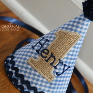 May include: A cone-shaped party hat featuring a light blue and white gingham pattern, topped with a dark blue yarn pom-pom. The hat displays a burlap number one appliqu&eacute; with the name Henry embroidered in dark blue thread and navy trim.