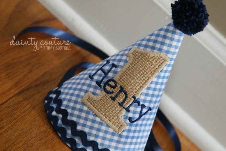 May include: A cone-shaped party hat featuring a light blue and white gingham pattern, topped with a dark blue yarn pom-pom. The hat displays a burlap number one appliqu&eacute; with the name Henry embroidered in dark blue thread and navy trim.