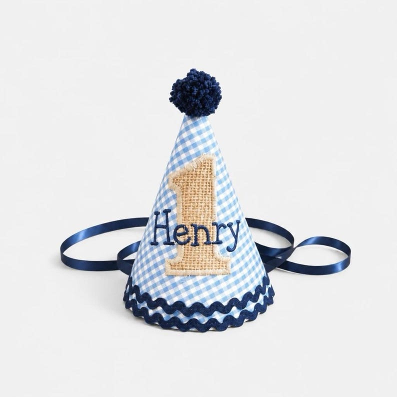 May include: A conical party hat featuring a light blue and white gingham pattern. The hat is topped with a dark blue pom-pom and trimmed with dark blue rickrack. A burlap number one is appliqued on the front with the name Henry embroidered in dark blue thread. Dark blue satin ribbons are attached to the sides.