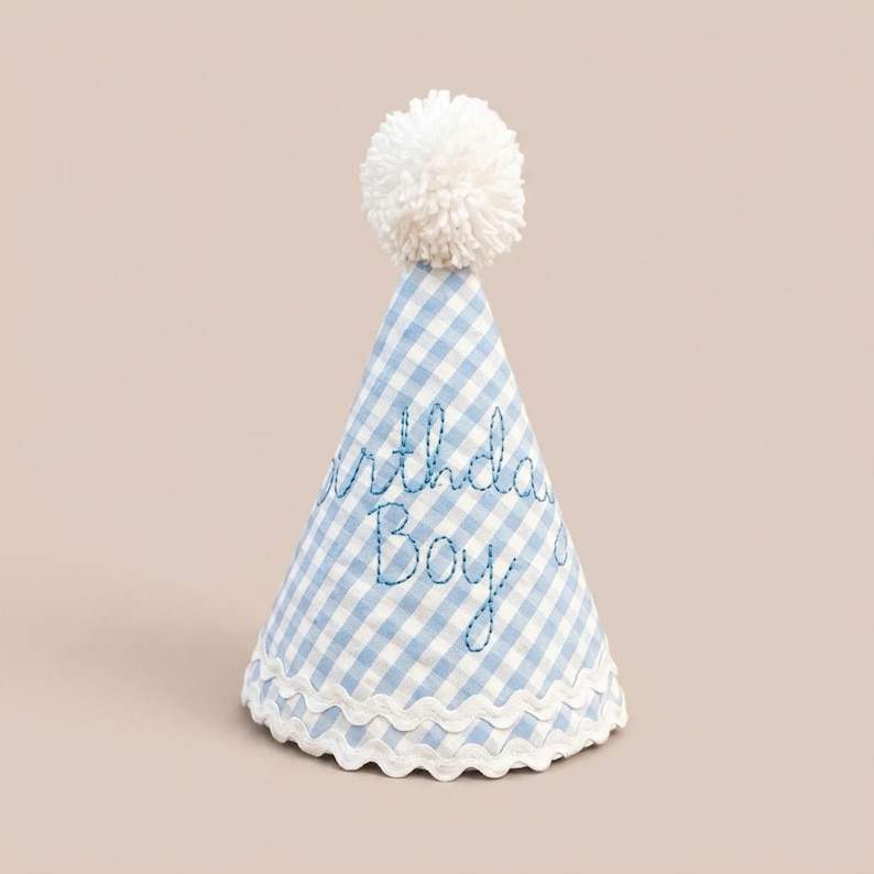 May include: A conical party hat featuring a light blue and white gingham pattern. The text "Birthday Boy" is embroidered in blue thread on the front. The hat is topped with a white yarn pom-pom and finished with a white scalloped trim at the base.