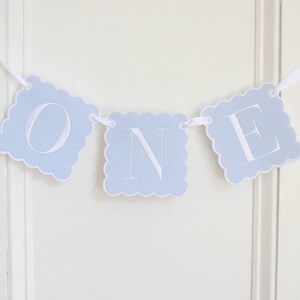 May include: A decorative banner featuring three light blue square cards with scalloped edges, each displaying a large white letter to spell out the word ONE. The cards are strung together on a thin white ribbon for hanging display.