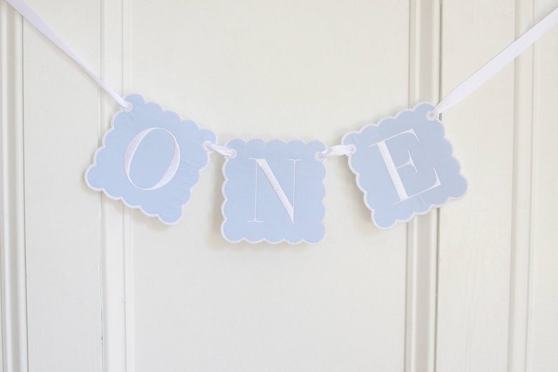 May include: A decorative banner featuring three light blue square cards with scalloped edges, each displaying a large white letter to spell out the word ONE. The cards are strung together on a thin white ribbon for hanging display.