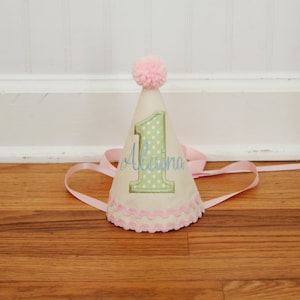 May include: A green and white polka dot party hat with a pink pom pom on top. The hat is embroidered with the number "1" and the name "Alaina".