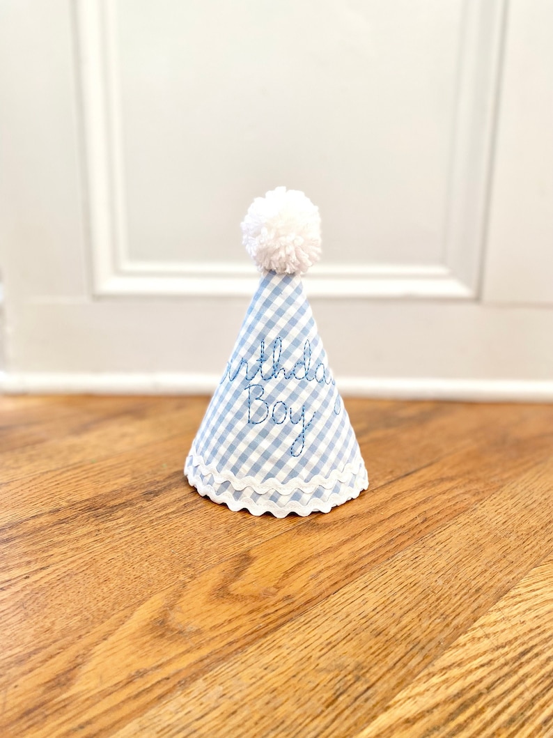 May include: A conical party hat featuring a light blue and white gingham pattern. The hat is topped with a white yarn pom-pom and finished with a white scalloped trim at the base. The text Birthday Boy is embroidered in blue thread on the front.