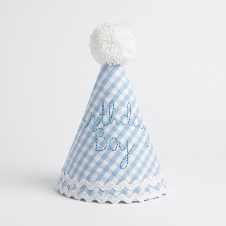 May include: A conical party hat featuring a light blue and white gingham pattern. The hat is topped with a white yarn pom-pom and trimmed with white rickrack at the base. The text "Birthday Boy" is embroidered in blue thread on the front.