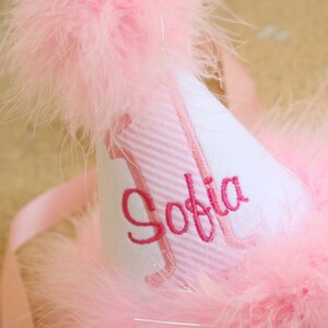 May include: A pink and white party hat with a pink and white striped band and the name "Sofia" embroidered in pink. The hat is trimmed with pink feathers.