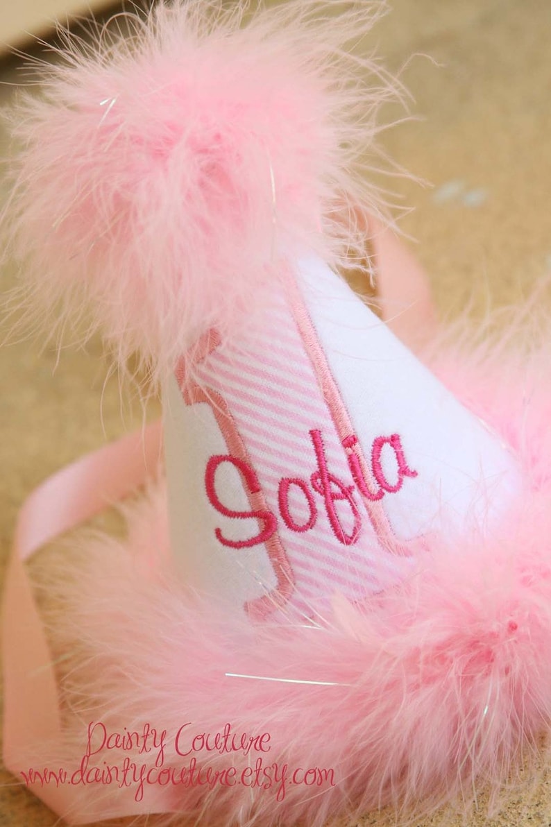 May include: A pink and white party hat with a pink and white striped band and the name "Sofia" embroidered in pink. The hat is trimmed with pink feathers.