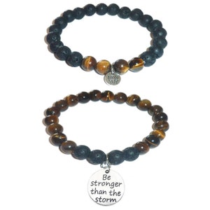 May include: Two beaded bracelets with charms. The top bracelet has black lava rock beads and tiger's eye beads with a charm that says "Om" The bottom bracelet has tiger's eye beads and black lava rock beads with a charm that says "Be stronger than the storm".