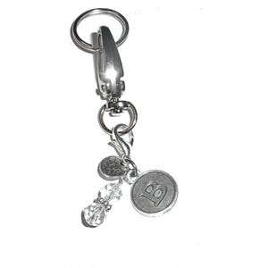 May include: A silver-toned metal keychain featuring a large lobster clasp and a split ring. Attached are three charms: a round disc embossed with the letter B, a small circular charm with a lotus flower design, and two clear faceted beads.