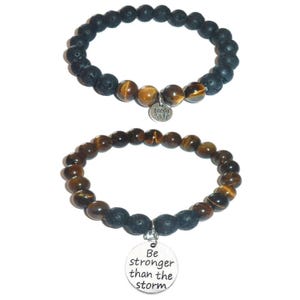 May include: Two beaded bracelets. The top bracelet features black porous lava beads and brown tiger eye beads with a silver lotus flower charm. The bottom bracelet consists of brown tiger eye beads and black lava beads with a round silver charm inscribed with the text Be stronger than the storm.