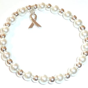 May include: A stretch bracelet featuring alternating white pearl-like beads and smaller metallic silver-toned spherical beads. A single silver-toned awareness ribbon charm hangs from the top of the circular beaded strand, centered on the bracelet.