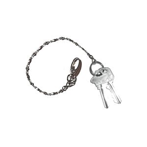 May include: A silver-colored key chain with a lobster clasp and a decorative chain. Two keys are attached to the chain. The keys are silver and have a simple design. The key chain is set against a white background.