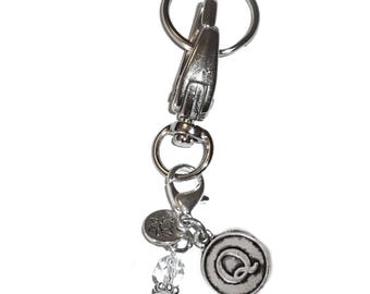 May include: A silver-toned metal keychain featuring a large lobster clasp and a split ring. Attached are three charms: a round metal disc embossed with the letter Q, a small circular charm with a lotus flower design, and two clear faceted beads.