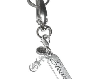 May include: A silver-toned metal keychain featuring a lobster clasp, a circular split ring, and two dangling charms. One charm is a clear, faceted bead, and the other is a rectangular metal tag engraved with the word Strong in cursive script.