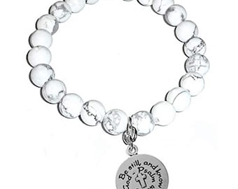 May include: A stretch bracelet composed of round white beads with grey veining. A circular silver-toned metal charm hangs from the beads, featuring a small cross and the engraved text: Be still and know that I am God - Psalm 46:10.