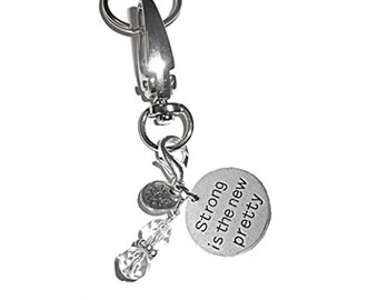May include: A silver-toned metal keychain featuring a lobster clasp and a split ring. Attached are a small circular charm, a clear faceted bead, and a larger round silver disc engraved with the text Strong is the new pretty in black lettering.