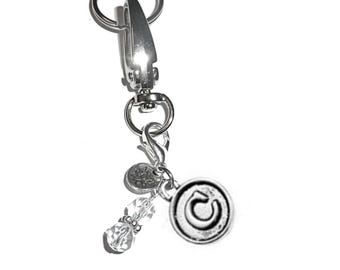 May include: A silver-toned metal keychain featuring a large lobster clasp and a split ring. Attached are three charms: a round silver disc embossed with the letter C, a small circular charm with a lotus flower design, and two clear faceted beads.