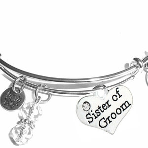May include: Silver expandable bangle bracelet with charms. The charms include a heart charm with the text "Sister of Groom", a crystal charm, and a charm with a lotus flower design.