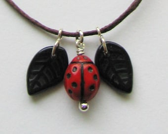 Puede incluir: Un collar de cordón marrón con un dije de mariquita roja y dos dijes de hojas negras.