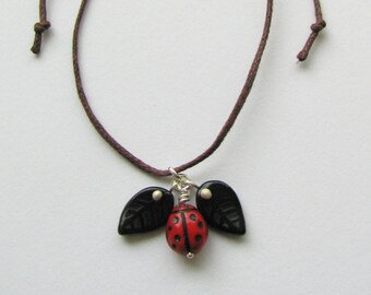 Puede incluir: Un collar de cordón marrón con un dije de mariquita y dos dijes de hojas negras. La mariquita es roja con manchas negras y las hojas tienen un punto blanco en cada una.