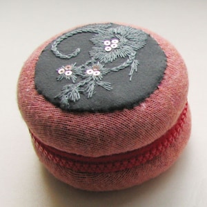 Peut inclure: Un coussin à épingles rond en tissu rose avec un motif floral brodé gris sur le dessus. La broderie est dans des tons de gris et comporte de petites paillettes. Une bordure tressée rouge accentue la base du coussin.