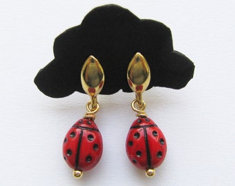 May include: A pair of gold ladybug earrings with red and black enamel. The earrings are hanging from a black display card.