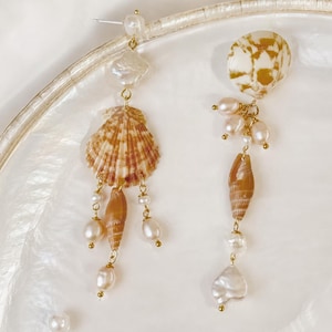 Op de afbeelding: Een paar asymmetrische oorbellen met natuurlijke schelpen, parels en goudkleurige accenten. Eén oorbel heeft een schelp met patroon en bungelende parels. De andere oorbel heeft een gespikkelde schelp en bungelende parels. Kustthema.