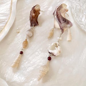 Op de afbeelding: Handgemaakte oorbellen met natuurlijke schelpen en kleine, donkerrode kralen. De oorbellen hebben een uniek, asymmetrisch ontwerp met een mix van schelpvormen en -maten, verbonden door fijne, zilverkleurige kettingen. De schelpen tonen een kleurenpalet van crème tot bruin.