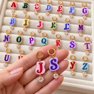 May include: A collection of gold-tone hoop earrings, each featuring a colorful, wavy-shaped acrylic letter charm and a small, clear, round cubic zirconia stone dangling at the bottom. The letters A through Z are displayed in various pastel and bold colors.