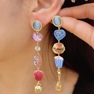 Puede incluir: Pendientes colgantes dorados con abalorios caprichosos. Un pendiente presenta una piedra azul, un corazón con texto, una gema transparente, un lazo, una frambuesa y una tetera. El otro tiene una piedra azul, un corazón, una hamburguesa, una magdalena y un tenedor.
