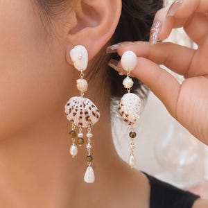 Op de afbeelding: Hangende oorbellen met natuurlijke schelpen, parels en kleine kralen. De oorbellen hebben een gelaagd ontwerp met een mix van texturen en kleuren, waaronder wit, bruin en goud. De oorbellen zijn ontworpen om op het oor te worden gedragen.