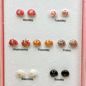 Può includere: Un set di sette paia di orecchini a lobo a forma di conchiglia, ognuno dei quali rappresenta un giorno della settimana. Gli orecchini presentano vari colori e motivi. La parola "JEWELLERY" è in alto e "Set G" in basso.
