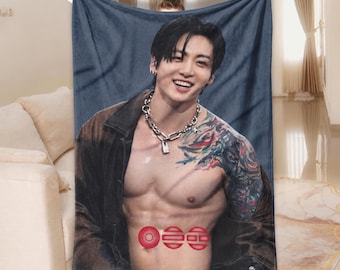 May include: A rectangular fleece blanket featuring a photographic print of a smiling person with dark hair, wearing a silver chain necklace and an open brown jacket. The person has visible tattoos on their arm and chest, with three red circular icons.
