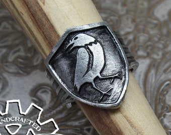 May include: A silver ring with a shield design featuring a detailed carving of two birds. The ring is handcrafted and has the words "Doctor Gus Original" engraved on the side.