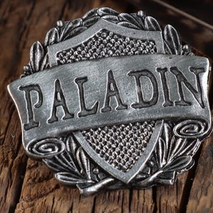 May include: A silver-toned metal pin featuring a shield design and the word "PALADIN" on a banner. The pin is displayed on a wooden surface.