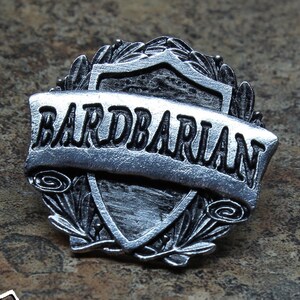 May include: A silver-toned metal pin with a shield design and the text "Bardbarian" on a banner. The pin is handcrafted and features a detailed design.