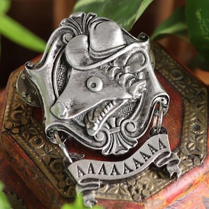 May include: A silver-colored metal pin featuring a rat wearing a hat, with a banner that reads "AAAAAAA". The pin is set on a decorative wooden box with a red and gold finish, surrounded by green plants.