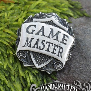 May include: A silver-toned metal pin with a scroll design and the words "GAME MASTER" on it. The pin is on a bed of green moss. The pin is handcrafted by Doctor Gus.