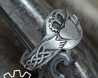 Może przedstawiać: Srebrny pierścionek Claddagh, z sercem, koroną i dłońmi. Pierścionek jest pokazany na srebrnej powierzchni. Obręcz pierścionka ma wzór celtyckiego węzła. Obraz zawiera tekst "HANDCRAFTED DOCTOR GUS ORIGINAL".