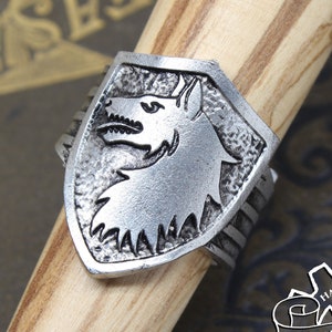 Op de afbeelding: Een zilveren ring met een wolfskopontwerp op een schild. De ring is handgemaakt en draagt de tekst "Doctor Gus Original".