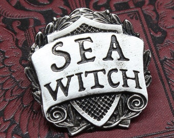 May include: A silver-toned metal pin featuring a shield-shaped design. The shield has a scroll with the words "SEA WITCH" on it. The pin is handcrafted by Doctor Gus.