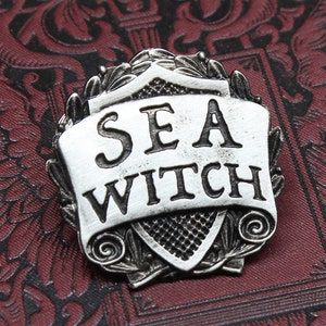 May include: A silver-toned metal pin with a shield-shaped design. The shield has a scroll with the words "SEA WITCH" on it. The pin is handcrafted by Doctor Gus.