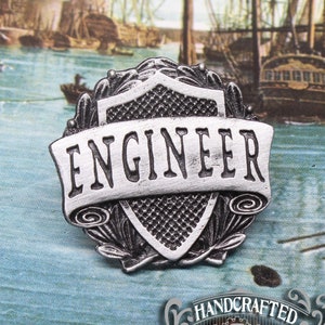 May include: Silver-toned metal pin with a shield design and the word "ENGINEER" in a banner. The pin is handcrafted and features a detailed design.