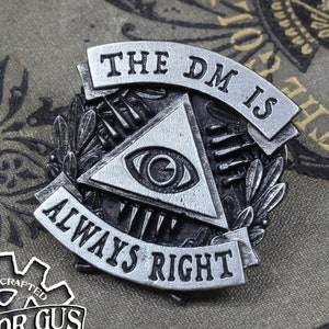 May include: A silver-toned pin with a black eye inside a triangle, surrounded by a wreath. The pin reads "The DM is Always Right". The pin is on a vintage book with a gold-colored design.